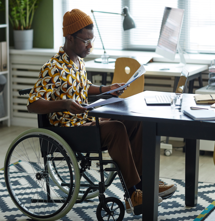 A man in a wheelchair, sitting at a desk with papers, working on documents.